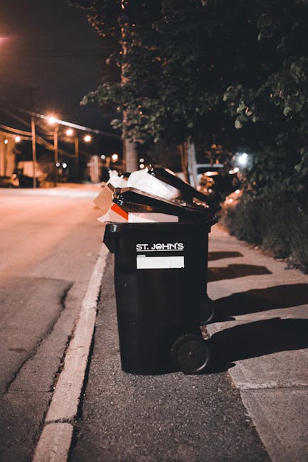 A black wheelie bin with the label 'ST. JOHN'S' in white text is positioned on the curbside of a dimly lit street during nighttime. The bin is filled with various rubbish, including cardboard boxes, paper, and plastic items protruding from the open lid. The bin’s surface appears slightly textured, and it has two small wheels visible at the base, which are partially shadowed. Behind the bin, there are dark, leafy trees with dense foliage casting shadows on the pavement. To the left, streetlights with warm yellow glow illuminate the road and pavement, with some lights visible in the distant background. The surrounding environment includes a concrete curb and a narrow sidewalk, which appears clean but slightly worn. The scene conveys a typical urban setting where private waste collection or bin servicing by a waste management company such as House Clearance Belgravia might take place, emphasizing the importance of proper rubbish disposal in residential areas, especially during evening hours when street lighting highlights the presence of waste bins awaiting collection.