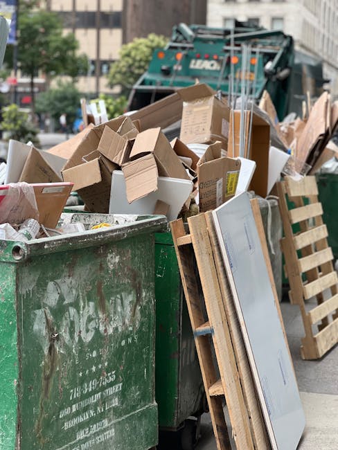 A large green waste container overflowing with various types of rubbish, including numerous cardboard boxes of different sizes, some crushed or partially flattened, scattered around and leaning against the sides. The boxes are made of brown corrugated cardboard, with some displaying printed labels or tape. To the right of the container, there is a wooden pallet standing vertically, supporting a flat, rectangular panel, possibly a piece of furniture or insulation material, with a smooth, light-colored surface. In the background, an urban street scene is visible with a construction or waste collection truck parked behind the container, partially obscured by the debris; the truck is dark green with visible equipment on top. The scene is outdoors, on a paved street with a few trees and multi-story buildings with windows and facades visible in the distance. The lighting appears natural, likely during daytime, highlighting the textures of the cardboard, wood, and metal. This scene illustrates a typical privately managed rubbish collection area, possibly part of an independent waste removal service, such as House Clearance Belgravia, handling bulk waste for a property clearance or refurbishment in an urban environment.