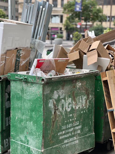A close-up view of a large, weathered green waste disposal container filled with various discarded materials, including cardboard boxes, folded and crushed, some with visible printing and tape, along with white and brown packaging materials. The container is situated outdoors on a paved surface, surrounded by other similar bins and a cluttered environment. In the background, there is an urban setting with buildings, trees, and a few parked vehicles, indicating a street or courtyard area. The scene suggests an instance of independent rubbish collection or on-site clearance, with the container visibly overloaded and used for disposing of bulky packaging and packaging debris, typical of private waste handling services. The container's rough, scratched finish and the assorted waste materials reflect an active rubbish removal process managed by a waste clearance company, such as House Clearance Belgravia, operating in the area. The overall lighting appears natural, with daylight illuminating the scene, emphasizing the textures and colors of the waste materials and the container.