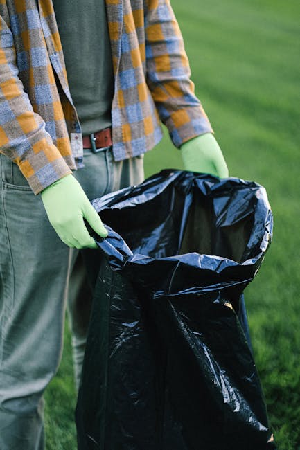 A person wearing a checked flannel shirt, grey trousers, and light green gloves is holding open a large black garbage bag in an outdoor setting with a grassy background. The bag appears to be made of shiny plastic material and is positioned upright, ready to be filled with waste or rubbish. The scene suggests environmental activity such as litter collection or waste disposal, which is relevant to rubbish removal services. The location looks like a park or garden area, with natural light illuminating the scene, emphasizing the texture of the bag and the uniformity of the gloves. The individual is facing away from the camera, and the focus of the image is on the open trash bag and the preparation for waste collection, aligning with the concept of independent or private disposal methods often involved in rubbish removal services like those offered by House Clearance Belgravia, maintaining a professional and neutral tone.