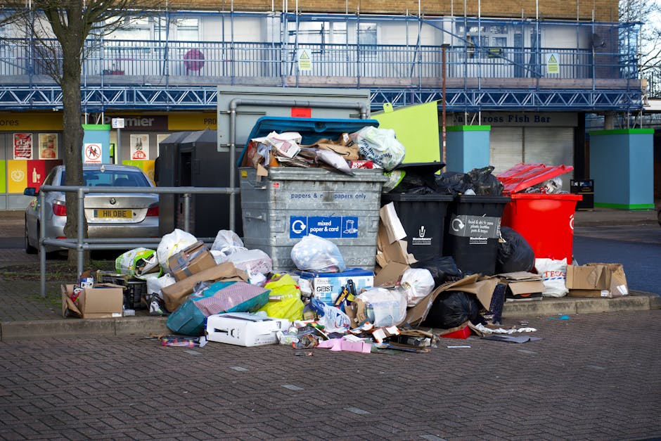 A large outdoor waste collection area located on a paved street, with an overflowing grey mixed paper and card waste container emitting various discarded papers, cardboard boxes, and packaging materials spread out in front of it. Several black and black plastic bags, some bulging with waste, are stacked next to the containers, along with a bright red recycling bin and additional cardboard boxes lying on the ground. Behind the waste area, there are storefronts with large glass windows and signage, and a building under construction or renovation covered in blue scaffolding. A parked silver car is situated adjacent to the waste collection point, separated by metal barriers, while leafless trees line the sidewalk. The scene is captured during daylight with overcast skies, highlighting the cluttered state of the rubbish and the surrounding urban environment, reflecting the need for professional rubbish removal services that handle private waste disposal efficiently and comprehensively.
