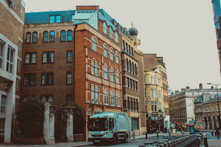 The image depicts a city street scene with several multi-storey buildings featuring varied architectural styles, including brick facades and ornate window frames. In the foreground, a small rubbish collection vehicle is parked adjacent to a sidewalk, with visible details of its light blue and white body, and a partially open rear hatch indicating recent loading activity. Surrounding the vehicle, there are bike racks and bollards along the pavement, with some greenery from small trees visible near a gated entrance on the left side. The background shows a mixture of historic and modern buildings, with a cloudy sky overhead casting diffuse light over the scene. The setting appears to be an urban area suitable for private waste collection services, such as those provided by House Clearance Belgravia, aligning with the context of residential and commercial rubbish clearance solutions. The overall environment is clean, with no visible rubbish or debris on the street or sidewalk.