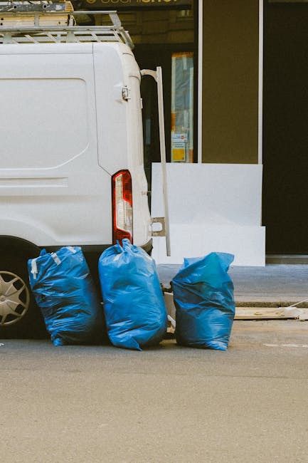 A narrow urban alleyway filled with large bags of mixed waste, including black plastic and fabric recycling sacks, positioned in the foreground alongside various debris and discarded items on the ground. In the background, there are densely stacked cardboard boxes and crumpled paper waste piled against a makeshift wooden barrier, indicative of private rubbish clearance efforts. The alley is bordered by graffiti-covered walls on the right and a weathered brick building with visible pipes and electrical fittings on the left. Leafless tree branches extend overhead, and the overall lighting suggests an overcast day or shaded environment, highlighting the gritty, cluttered atmosphere typical of an area awaiting rubbish removal or clearance services. This scene, associated with independent waste handling, underscores the importance of professional rubbish removal solutions like those provided by House Clearance Belgravia, especially in tight urban spaces where clutter accumulates.